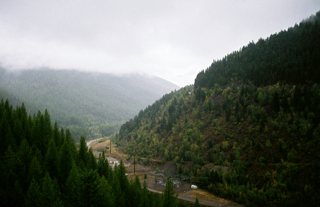 [The river runs straight for a short distance before heading right and out of view behind the huge hillside. The hillsides on either side of the river are steep and evergreen covered. There is a lot of fog at the skyline level on the left side.]